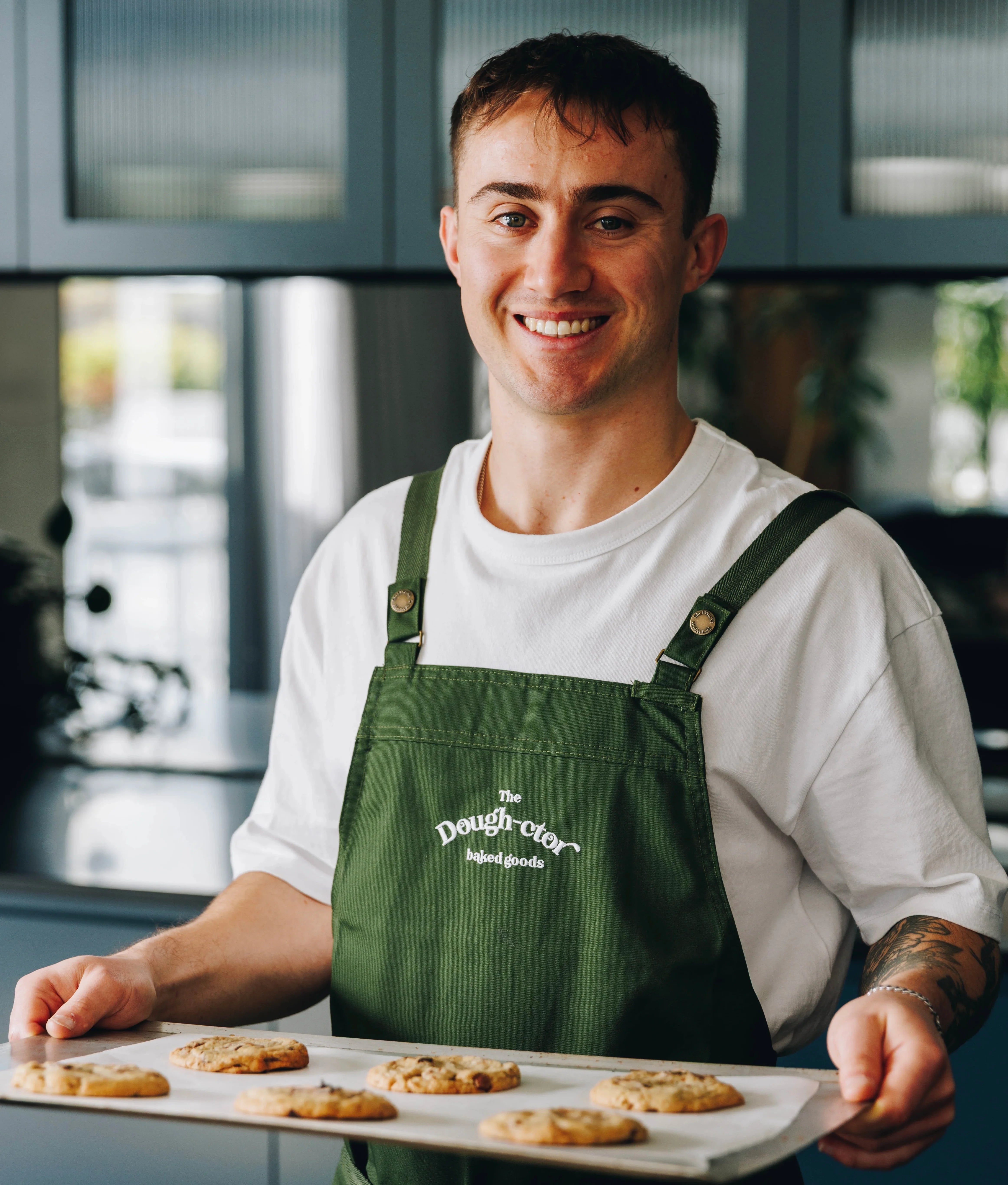 Man in a green apron holding a tray of cookies in a kitchen setting
