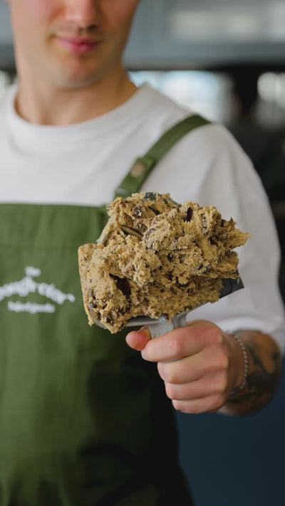 Man in green apron holding stand mixer paddle covered in cookie dough